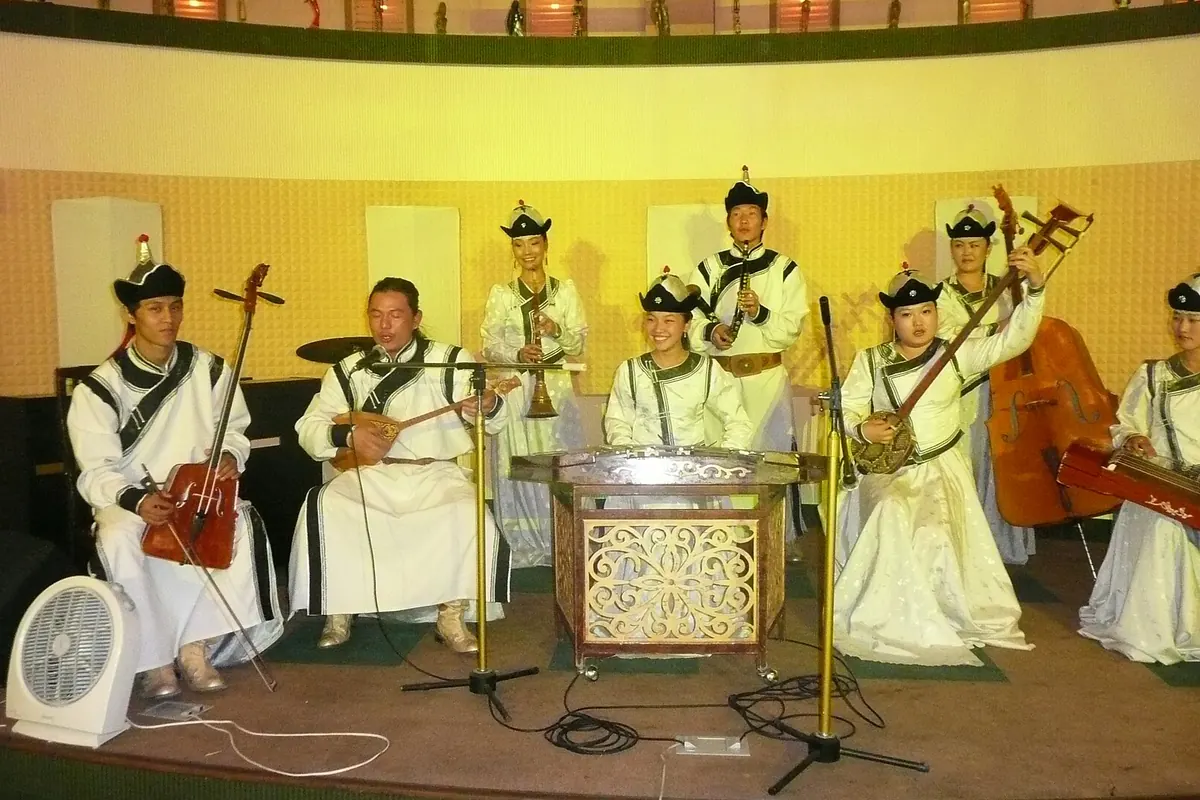 Mongolský folklorní soubor v bílých hedvábných kostýmech a černých kloboucích na malém pódiu, s houslemi, loutnou, cimbálem, citherem, dechovými nástroji a kontrabasem